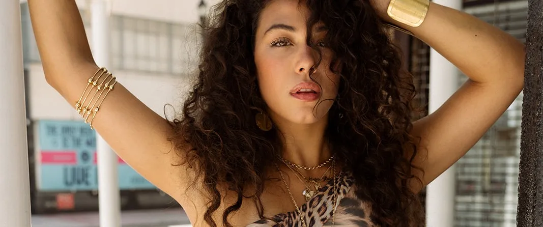 Bella Valdes looks directly into the camera, arms raised, dark curls loose, in a leopard-print top against sunlit downtown Miami architecture.