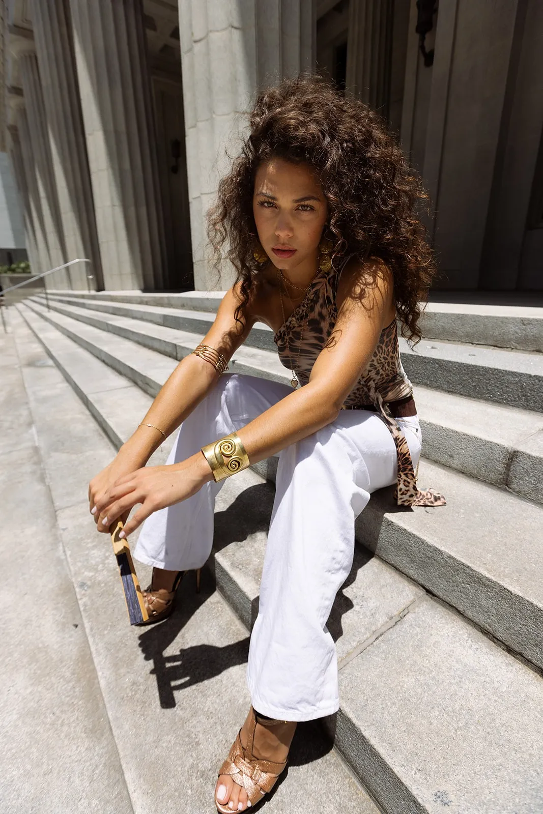 Bella Valdes seated on stone courthouse steps in white trousers and a leopard-print top, elbows on knees, looking into the camera.
